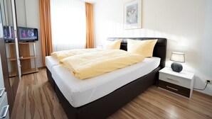 Une chambre d’hôtel avec un lit, une table de chevet avec une lampe, un téléviseur posé sur un meuble et une fenêtre avec des rideaux.
