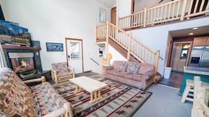 A cozy living room with a fireplace, a sofa, a chair, and a coffee table. There is a staircase leading to an upper level. - Continental Divide Cabin with breathtaking views from large picture windows (Estes Park)