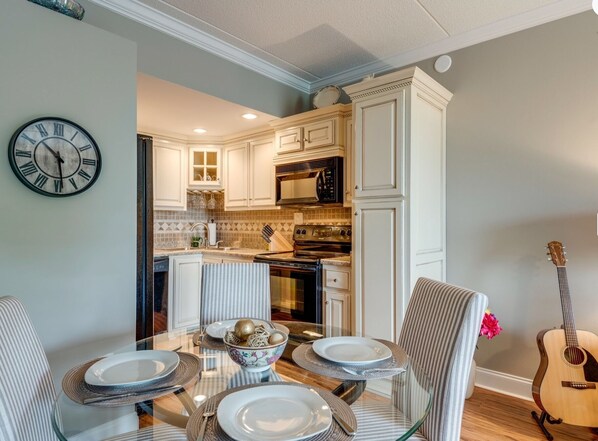 Un coin repas avec une table en verre dressée pour quatre personnes, entourée de chaises à rayures. Une horloge est fixée au mur, à gauche.