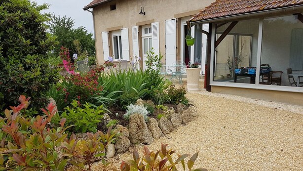 A house with a gravel driveway, a garden with various plants, and a dining area with outdoor furniture. - Renovated countryside family cottage with garden (Cindré)