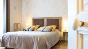A neatly made bed with a grey bedspread and multiple pillows, a wooden nightstand with a lamp, and a patterned wallpaper on the wall. - Renovated countryside family cottage with garden (Cindré)