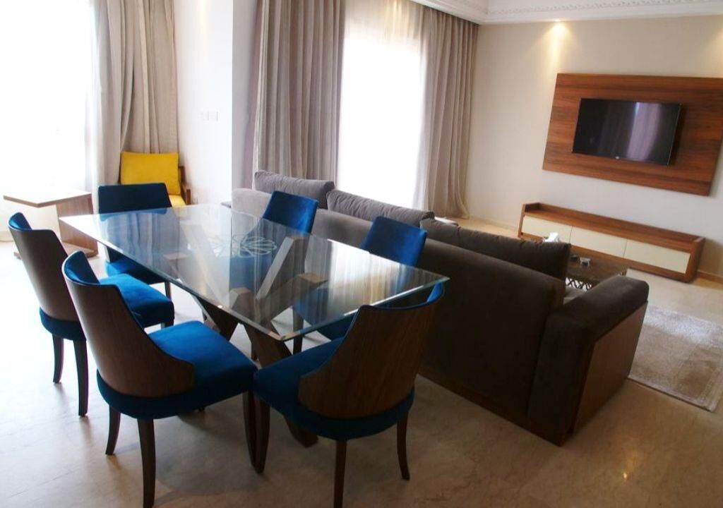 A modern living room with a glass-top dining table surrounded by blue chairs, a brown sofa, and a television mounted on the wall.