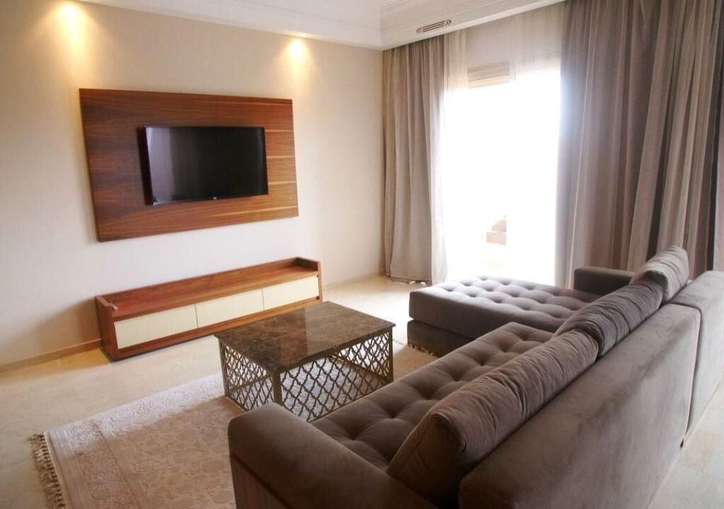Una sala de estar moderna con un televisor de pantalla plana montado en la pared, un sofá, una mesa de centro y un ventanal con cortinas.