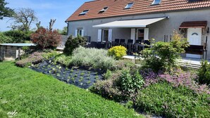 A two-story house with a tiled roof, a covered patio with outdoor furniture, and a garden with various plants and flowers. - Renovated farmhouse with private pool and pets allowed (Échassières)