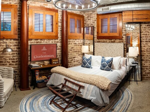 A cozy bedroom with a brick wall, wooden shutters, a bed with white bedding, and a vintage radio. - Charleston Studio | Steps to King, Private Suite & Patio | The Charlestonian (Charleston)