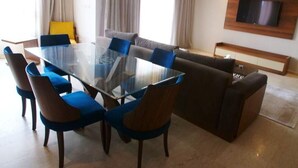 Een moderne woonkamer met een eettafel met glazen blad, omringd door blauwe stoelen, een bruine bank en een aan de muur bevestigde televisie.