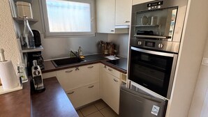 A compact kitchen with a built-in oven, microwave, and refrigerator. There is a window providing natural light and a small countertop with various kitchen items.