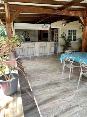 Een rustiek houten terras met een bar, witte barkrukken en een tafel met een blauwe tafelkleed.