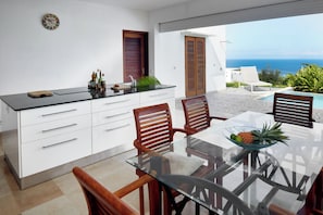 A modern kitchen with a glass table and wooden chairs, a view of the sea, and a pool area. - Villa Savane Paille 3. 2 (Deshaies)