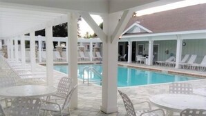 A covered pool area with lounge chairs and tables, a clear pool, and a building with a sloped roof in the background. - Close to Fun, Villa In Walking distance to Lake Sumter Landing w/Golf Cart! (The Villages)