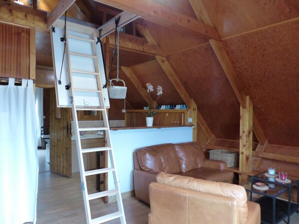 A cozy attic room with a wooden ceiling, a brown leather sofa, a ladder, and a small table with cups.