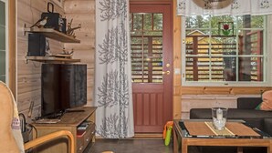 A cozy cabin interior with a wooden interior, a television on a wooden stand, a dining table with chairs, and a window with a view of a wooden fence and greenery outside.