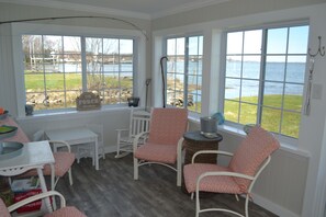 Une pièce confortable avec vue sur l’eau, comprenant des fauteuils en osier blancs, une petite table et une fenêtre donnant sur la plage.