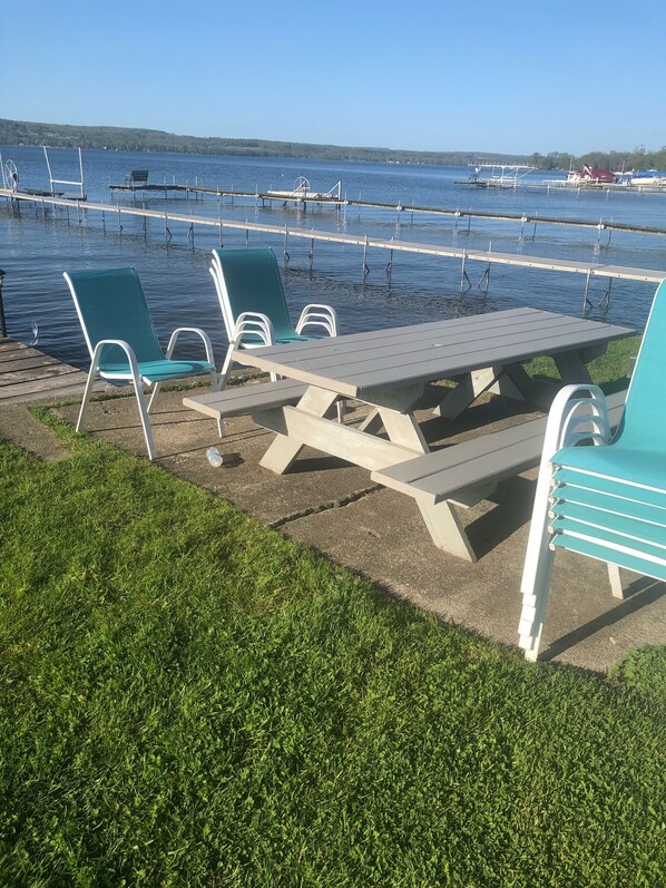 A lakeside picnic area with a table and chairs, a grassy foreground, and a dock with boats in the background. - 3 Bedroom Cottage on the Shores of Chautauqua Lake (Lakewood)