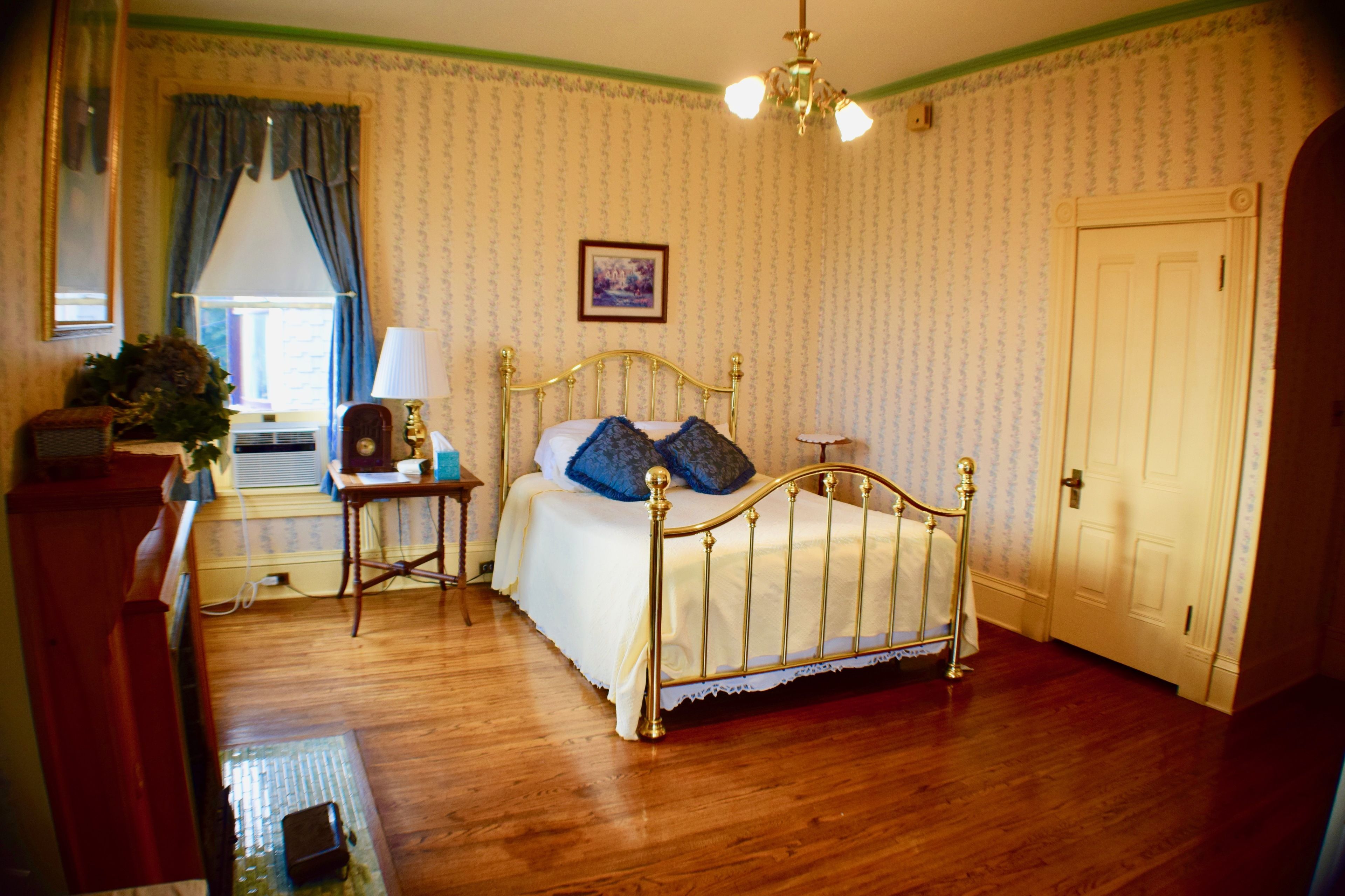 A bedroom with a wooden floor, a bed with a gold frame, a nightstand, a lamp, and a window with curtains.