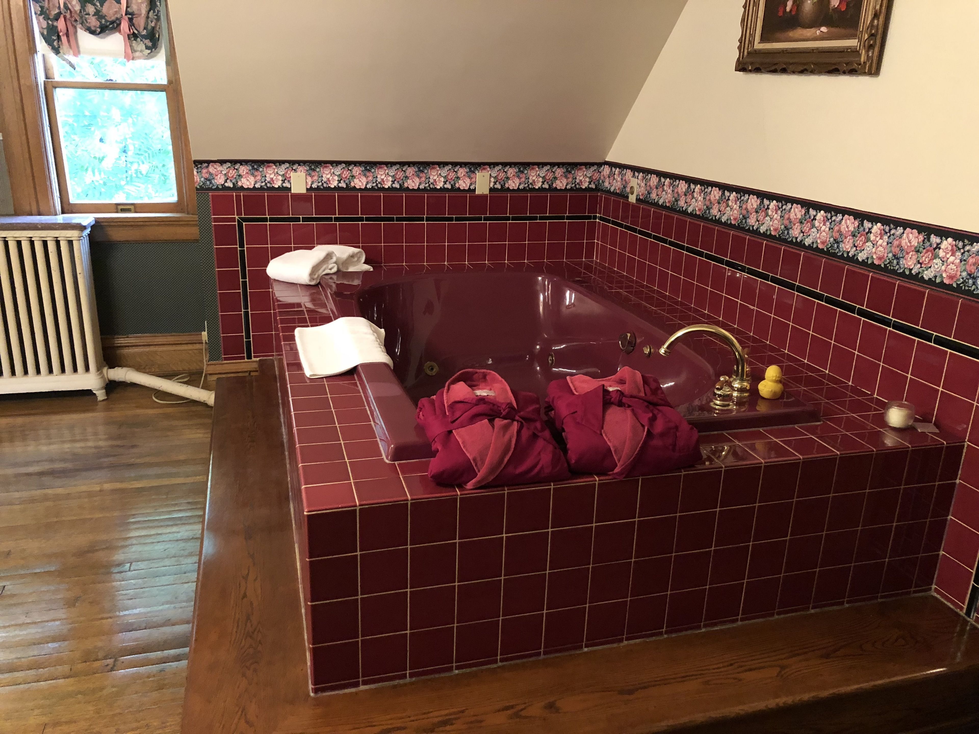 A bathroom with a red-tiled bathtub, a wooden radiator, and a window with floral curtains.
