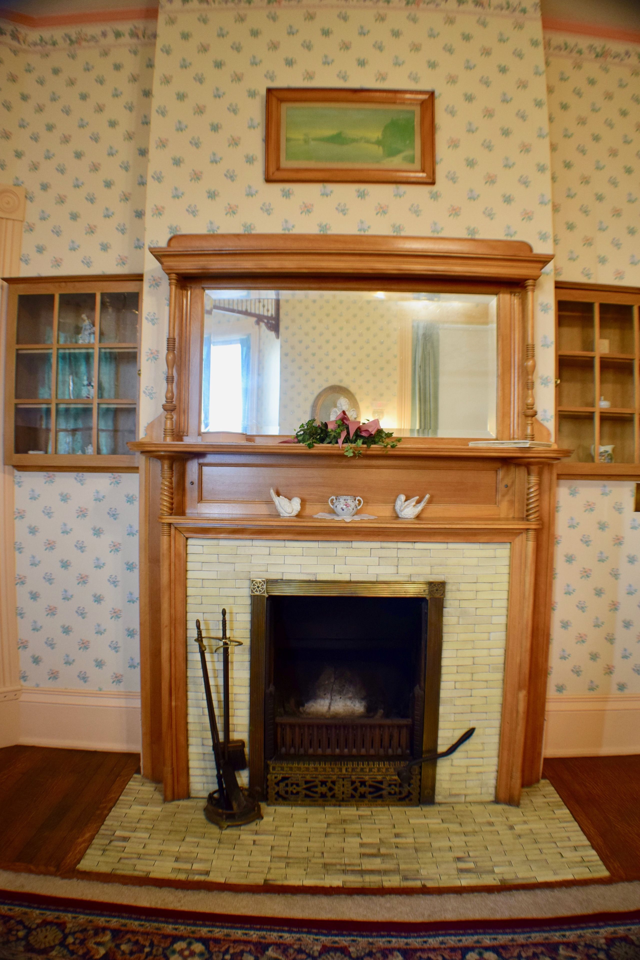 A fireplace with a wooden mantel, a mirror above it, and a floral wallpaper pattern on the walls.
