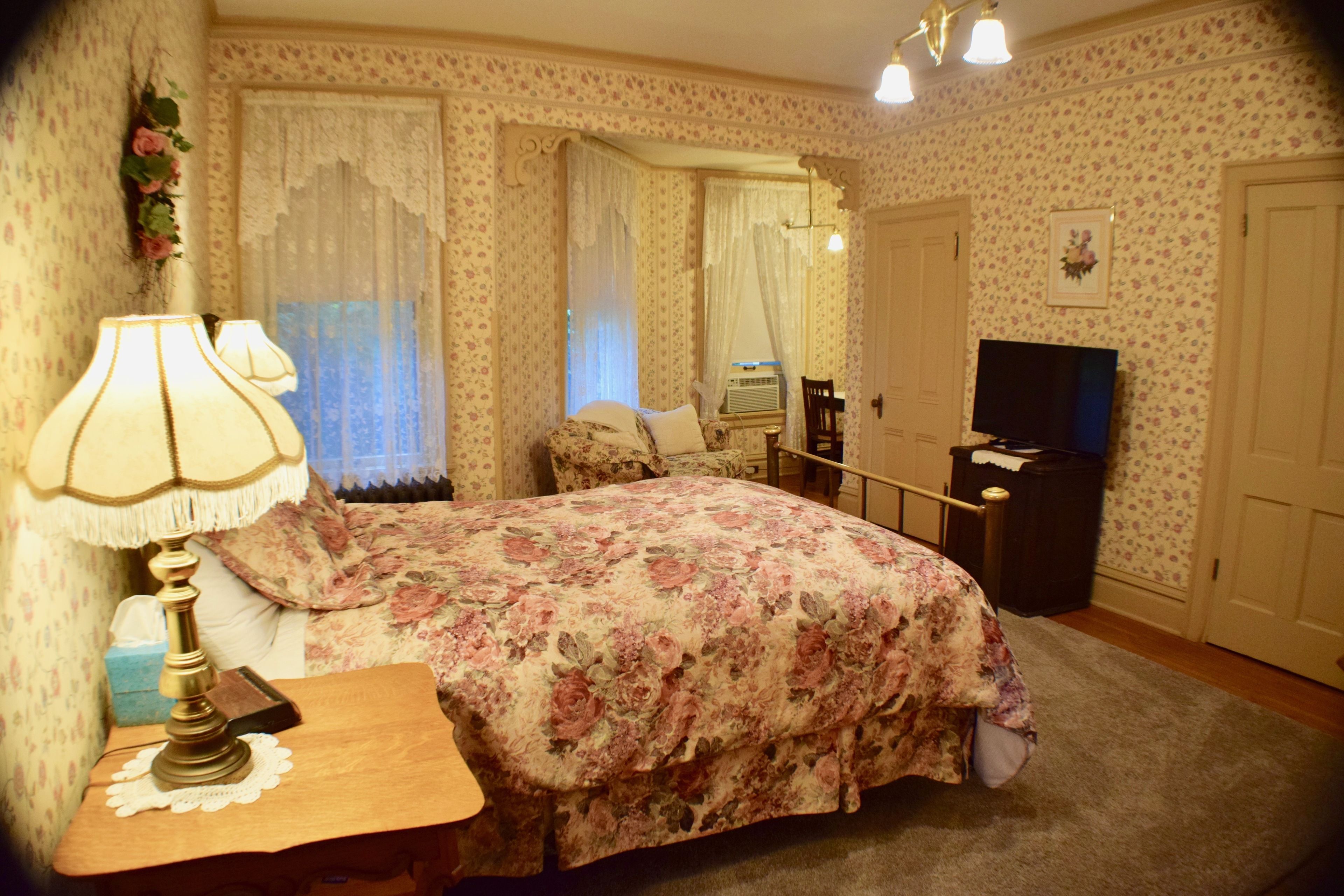 Quarto com colcha florida, abajur, cômoda, televisão e espelho.