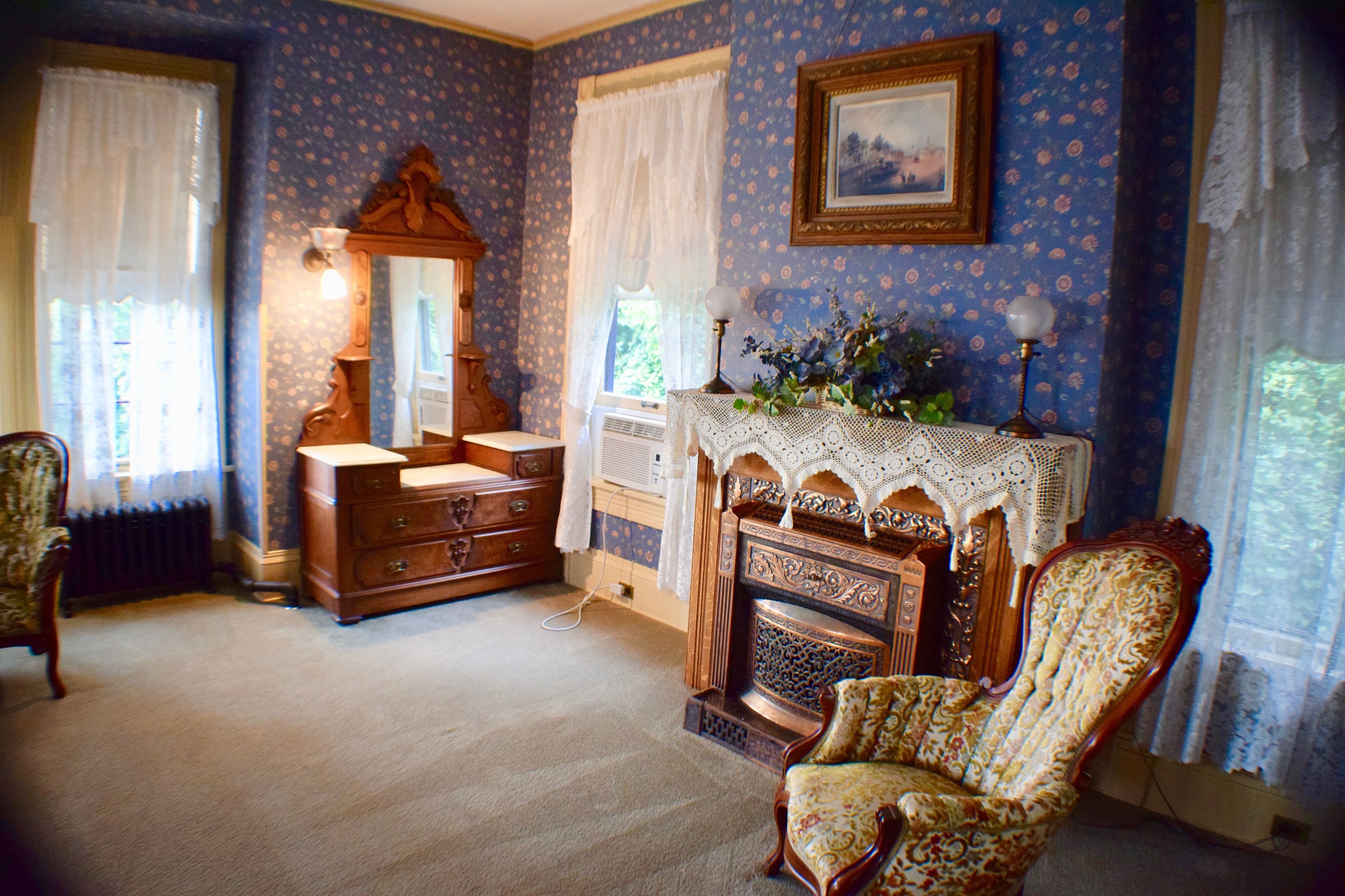 Une pièce comprenant une commode en bois, une chaise, une cheminée ornée d’une cheminée en dentelle et un tableau encadré accroché au mur.