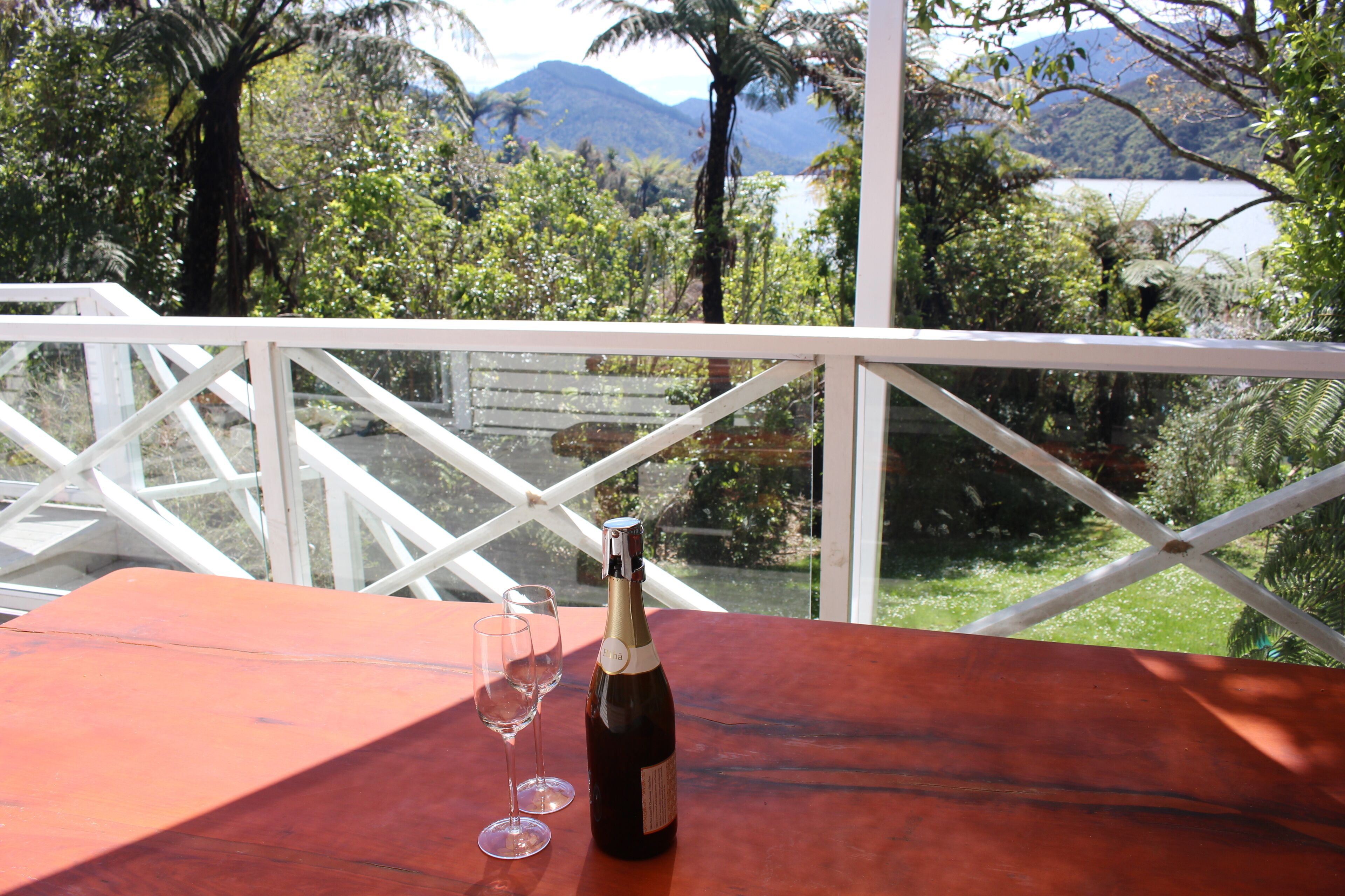 A wooden table with a bottle of wine and two glasses on a balcony overlooking a lush landscape with trees and a body of water.