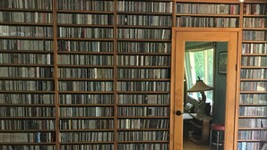 Une pièce dotée d’une grande bibliothèque remplie de livres et de DVD. On y trouve une porte en bois avec un miroir qui reflète une partie de la pièce.