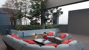 Un espacio moderno para sentarse al aire libre con mobiliario de mimbre y cojines rojos, rodeado de follaje y vistas a la ciudad.