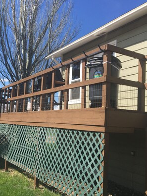 Una terraza de madera con un cerramiento de celosía, una ventana con reja y un árbol al fondo.