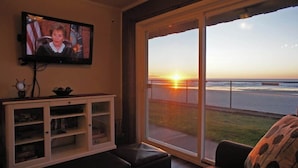 Un salon avec un téléviseur, un meuble blanc et une grande fenêtre donnant sur le coucher de soleil sur l’océan.