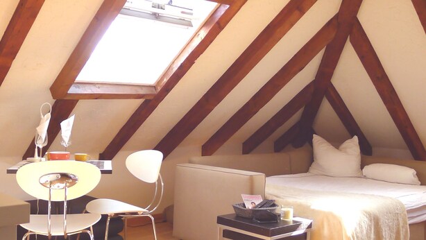 Ein Schlafzimmer im Dachgeschoss mit schrägem Dach, Holzbalken, einem Bett, einem Schreibtisch und einem Stuhl.