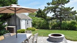 An outdoor patio with a table and chairs, a stone fire pit, and a wooden shed in the background.