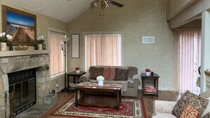 Ein geräumiges Wohnzimmer mit Kamin, Deckenventilator und großem Fenster mit Jalousien.