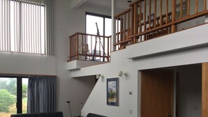 A spacious living area with a high ceiling, a balcony with wooden railings, a black leather sofa, and a glass coffee table. - Ocean View Matunuck Home - Go to Water, Go to the Village! (Wakefield)