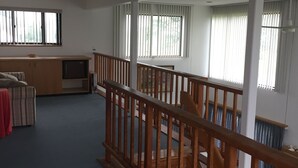 A spacious room with a wooden staircase, large windows with blinds, a sofa, and a television. - Ocean View Matunuck Home - Go to Water, Go to the Village! (Wakefield)