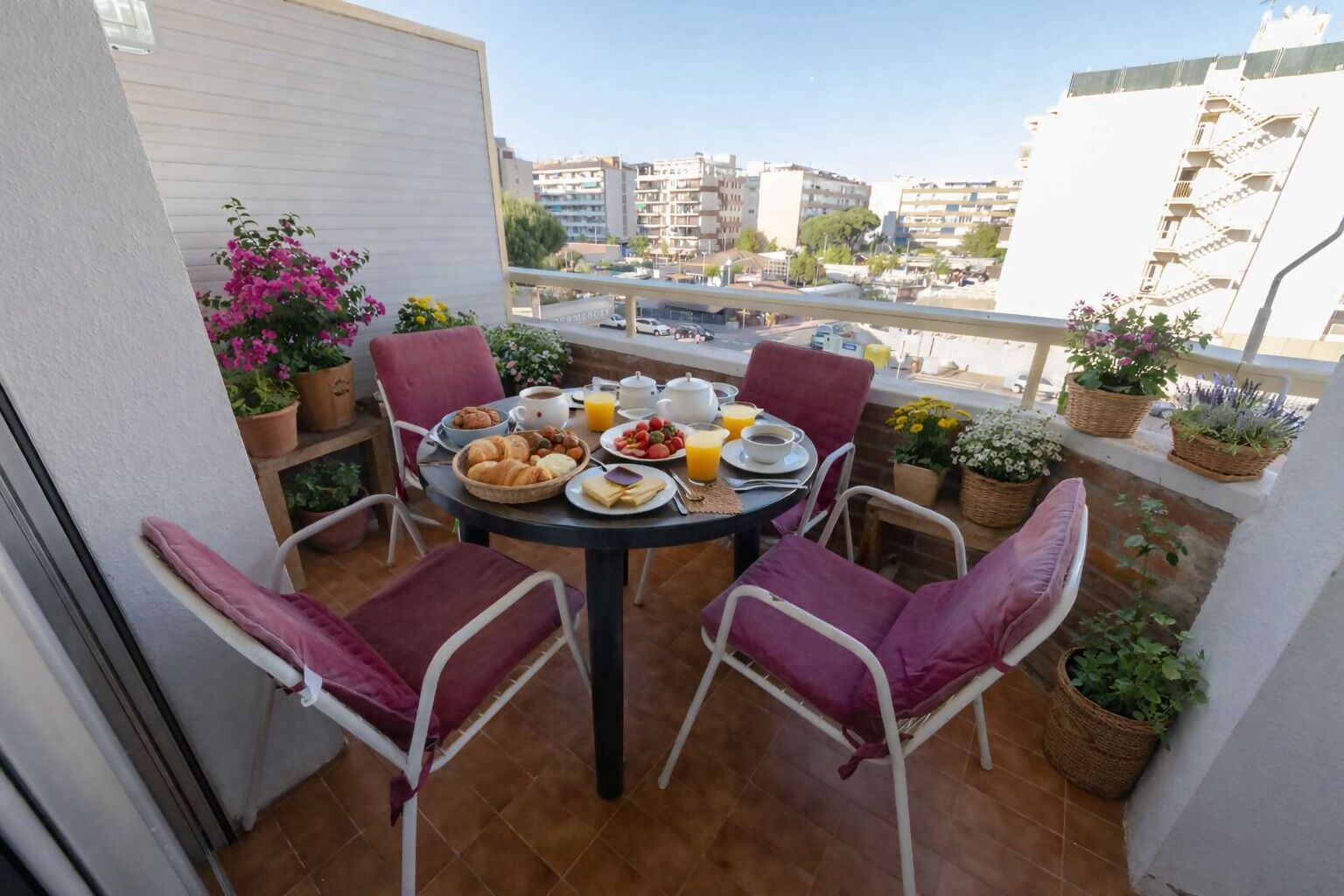 A balcony with a round table set for breakfast, surrounded by chairs, with potted plants and a city view.