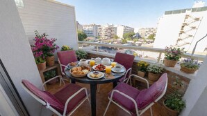 Un balcón con una mesa redonda dispuesta para el desayuno, rodeada de sillas, plantas en macetas y vistas a la ciudad.