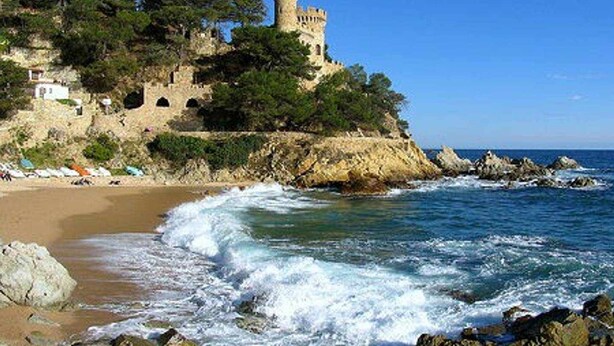Paisaje costero con un castillo en un acantilado, playa y costa rocosa.