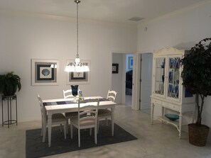 Un comedor con una mesa y sillas blancas, una araña, obras de arte enmarcadas y un aparador con puertas de cristal.