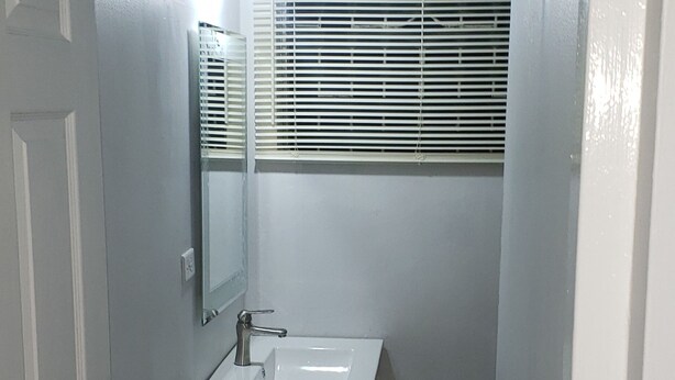 A narrow bathroom with a white sink, a mirror, and a window with blinds.
