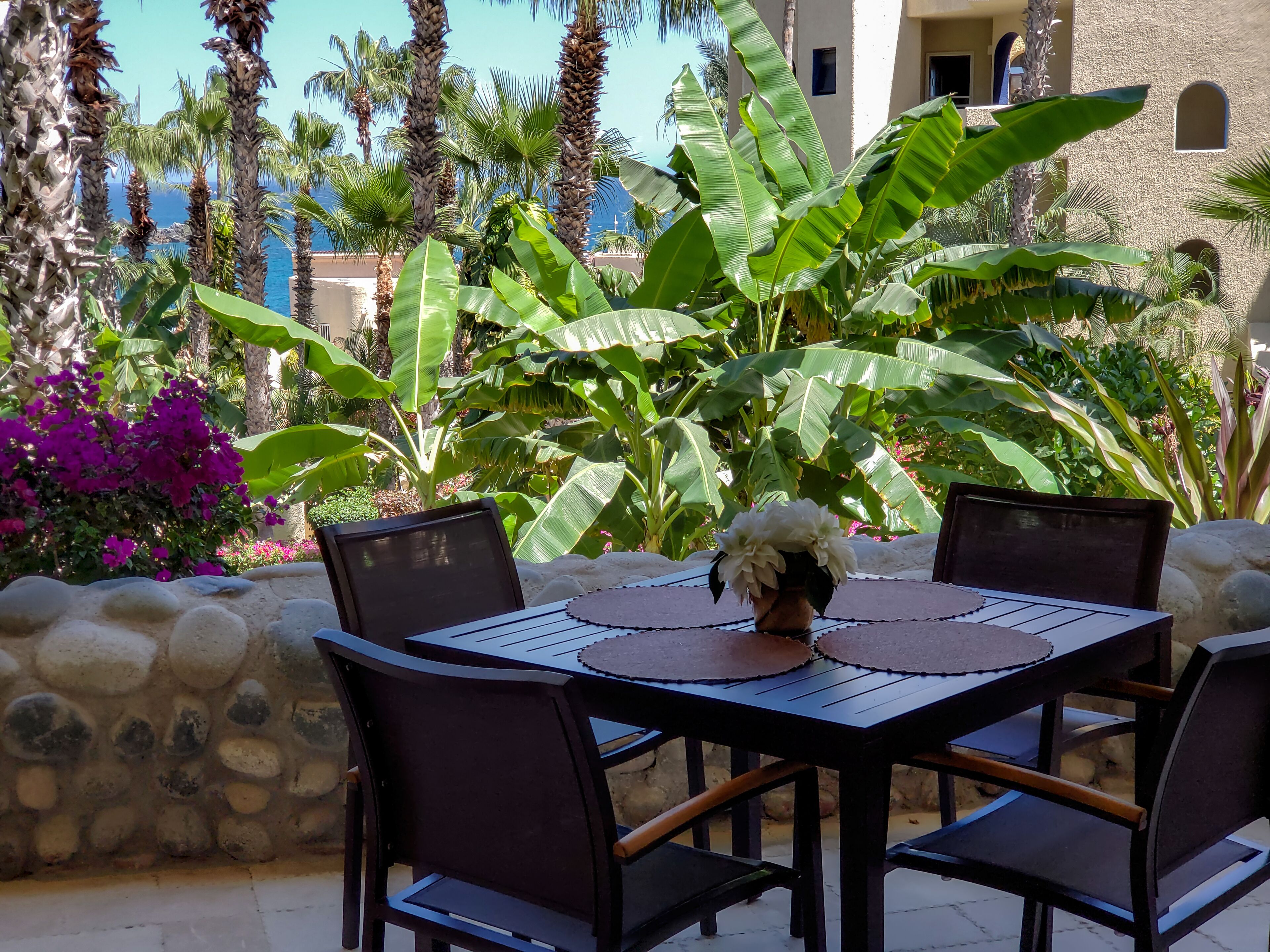 An outdoor dining area with a table and chairs, surrounded by tropical plants and a view of the ocean.