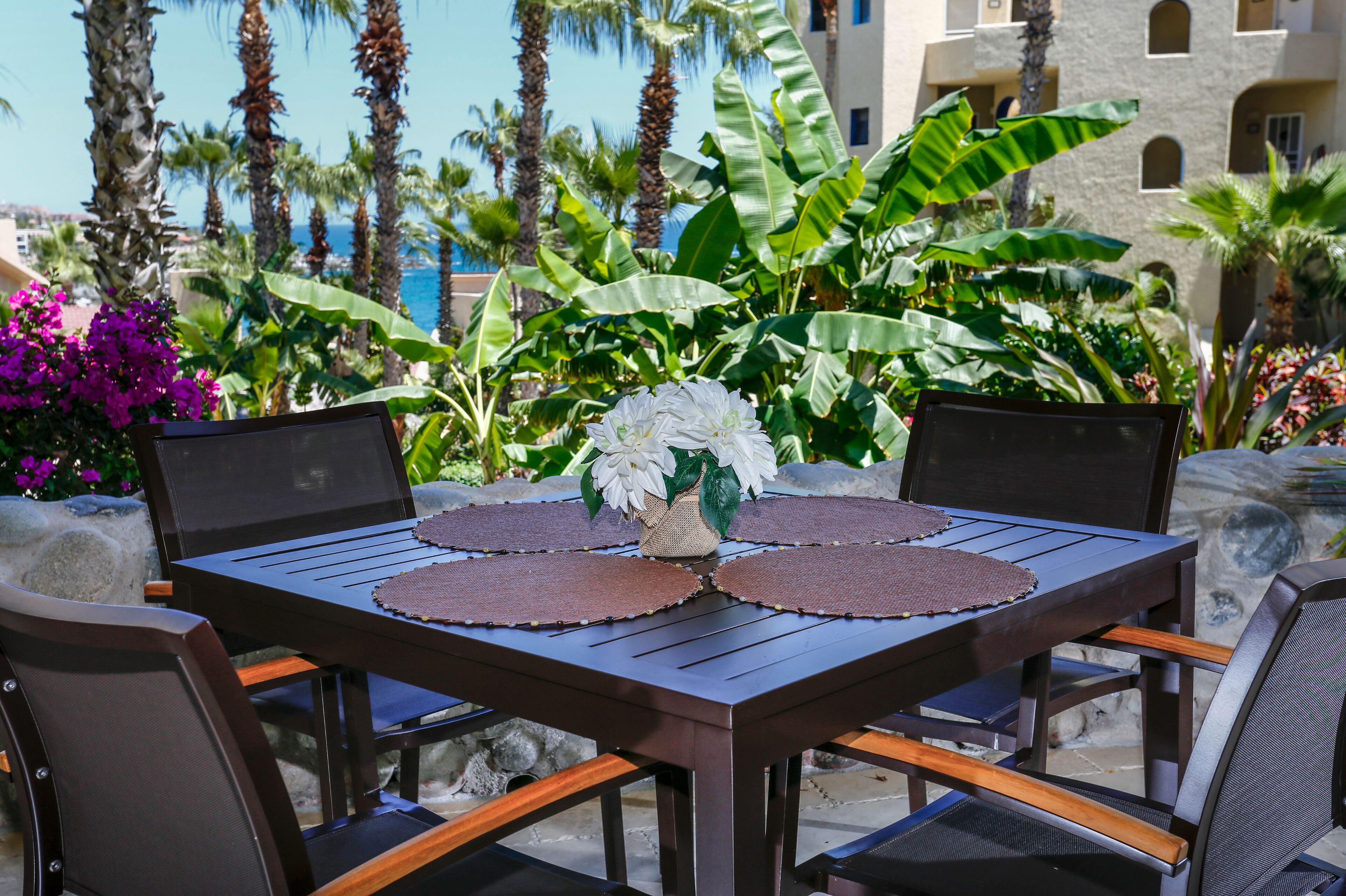 A wooden table with a vase of white flowers and a placemat, surrounded by chairs, set against a backdrop of palm trees and a building.