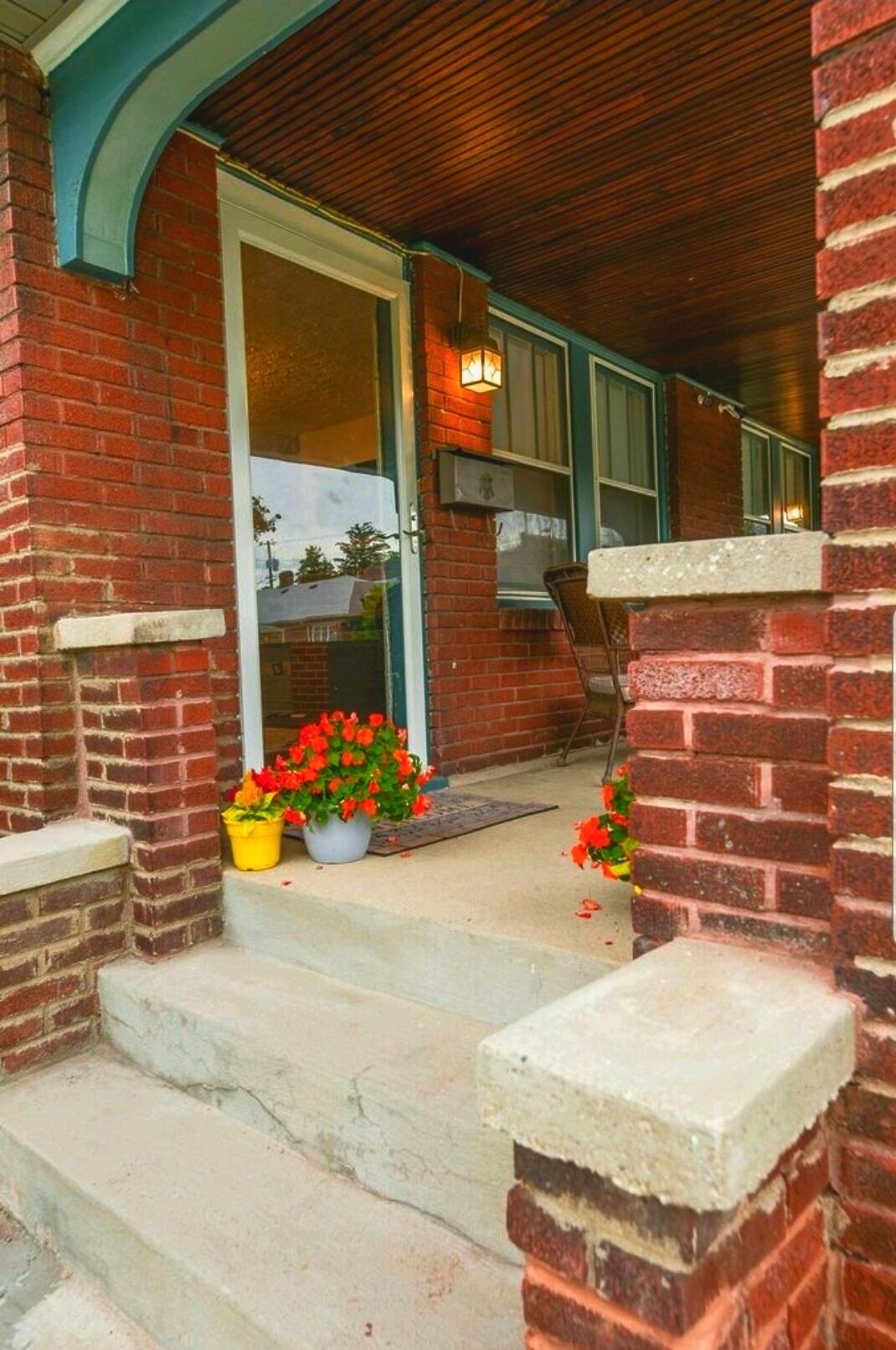 A brick house with a wooden porch, a door, and steps leading to the entrance. There are potted plants with red flowers on the steps.