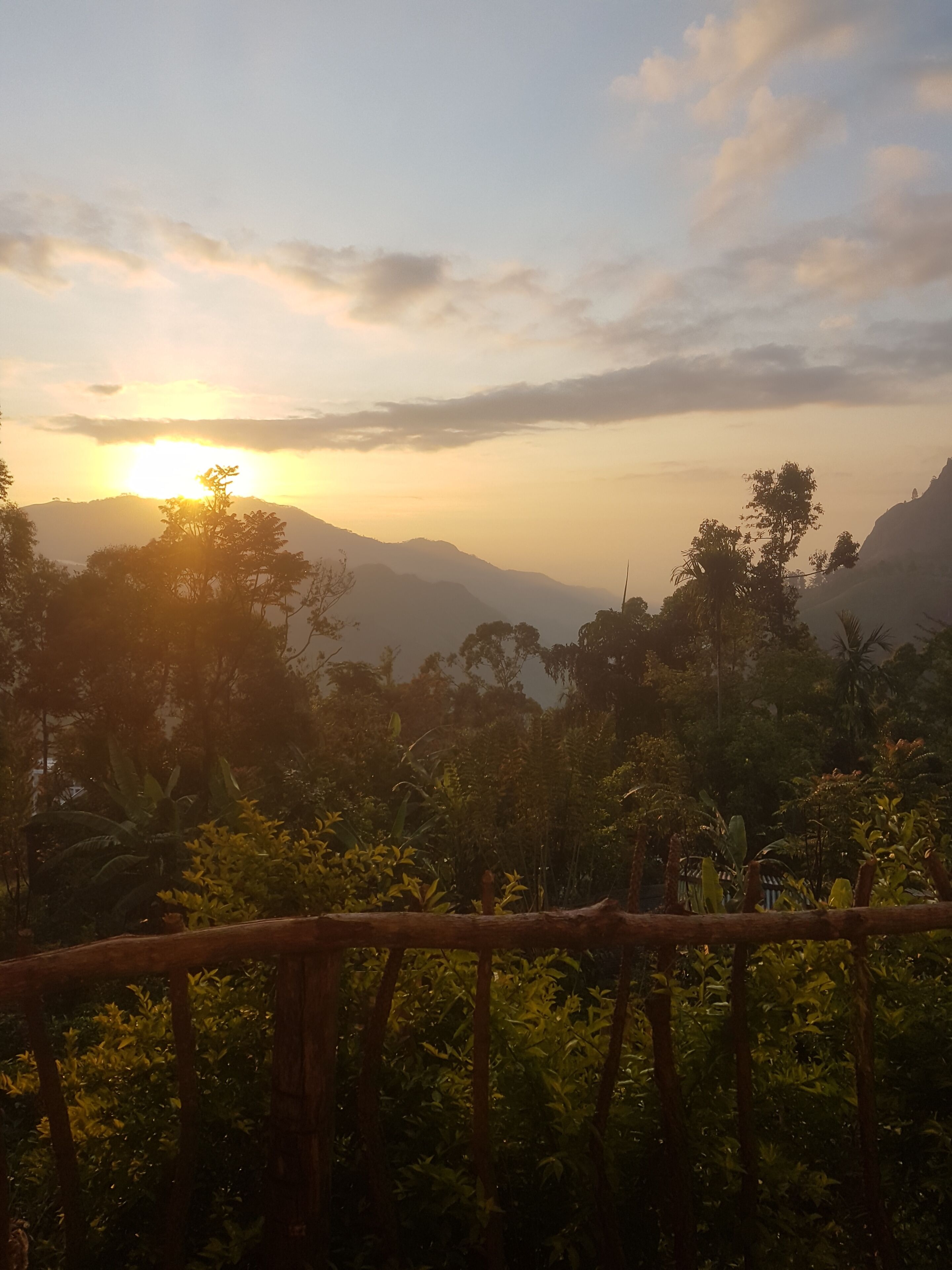 A sunset view from a balcony with a wooden railing, overlooking a lush landscape with mountains and dense vegetation.