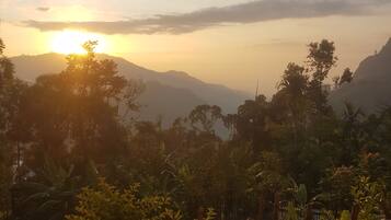 A sunset view from a balcony with a wooden railing, overlooking a lush landscape with mountains and dense vegetation.