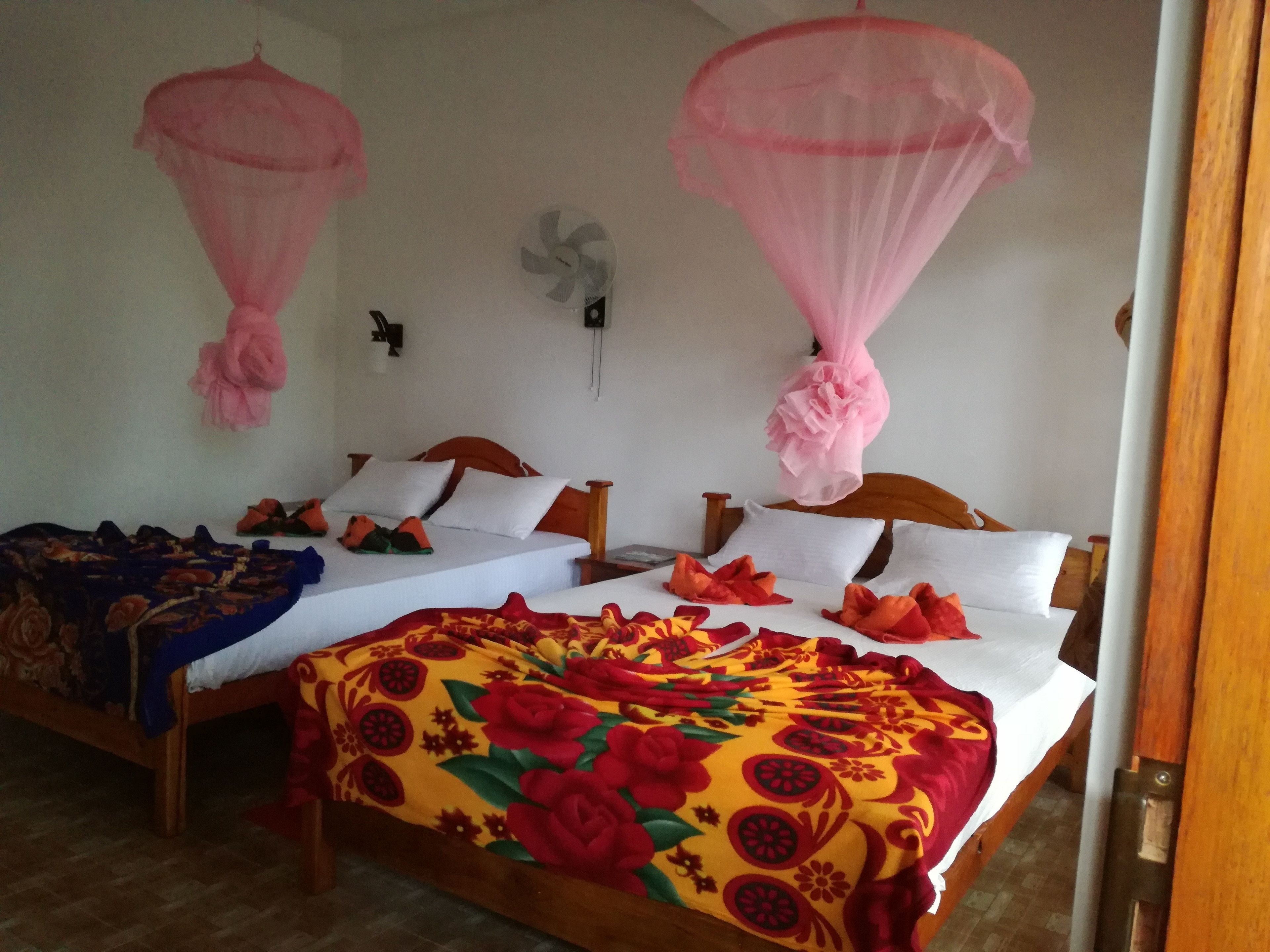 Ein Zimmer mit zwei Betten, jedes mit Baldachin. Es gibt einen Wandventilator und eine Wandleuchte.