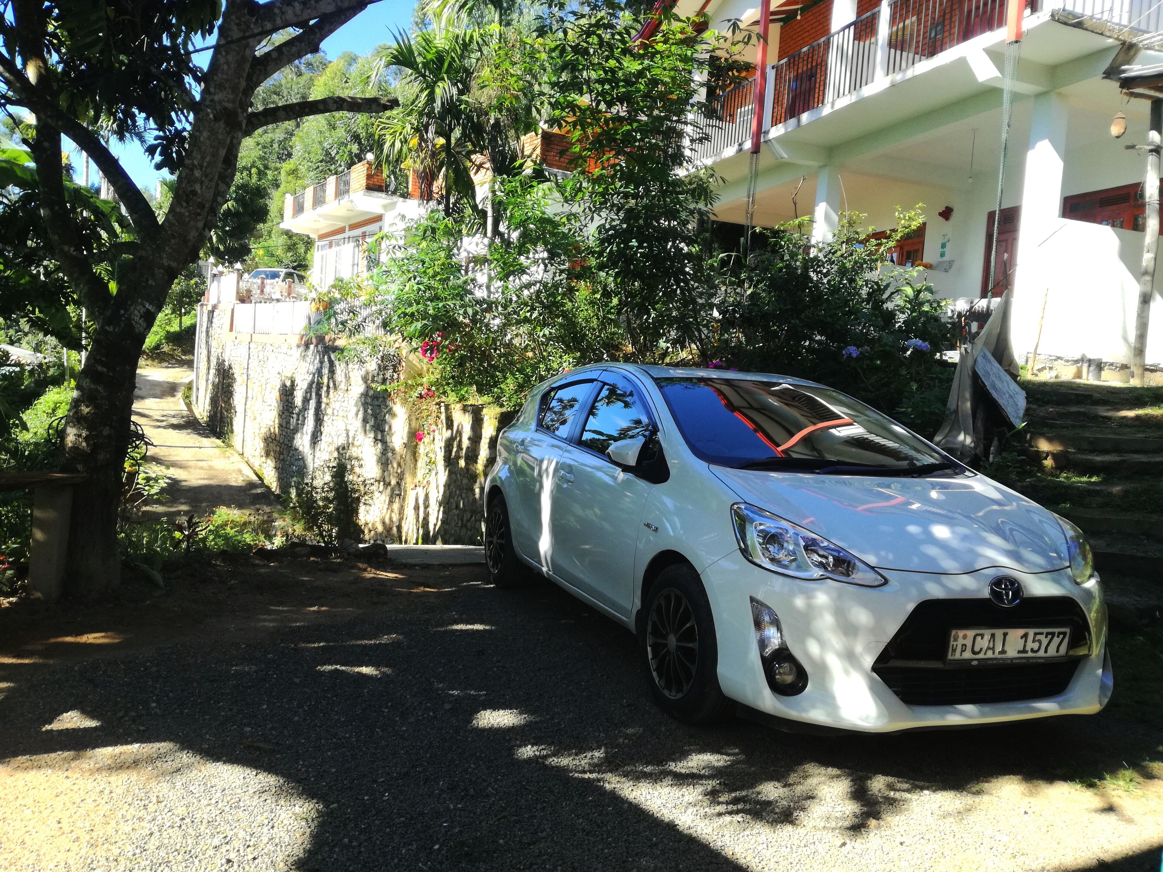 A white car parked on a gravel path with a stone wall and greenery on one side, and a building with balconies on the other.