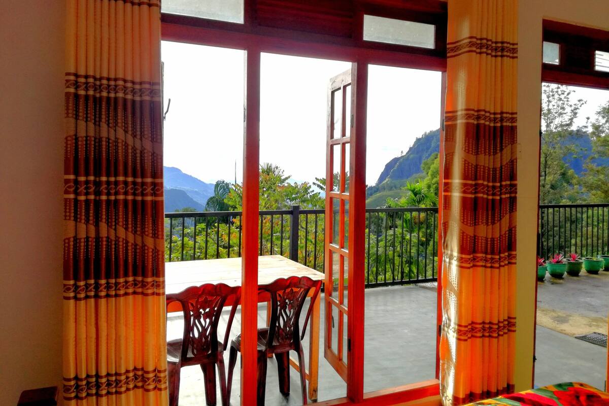 Una habitación con mesa y sillas, un balcón con barandilla y vistas a montañas.