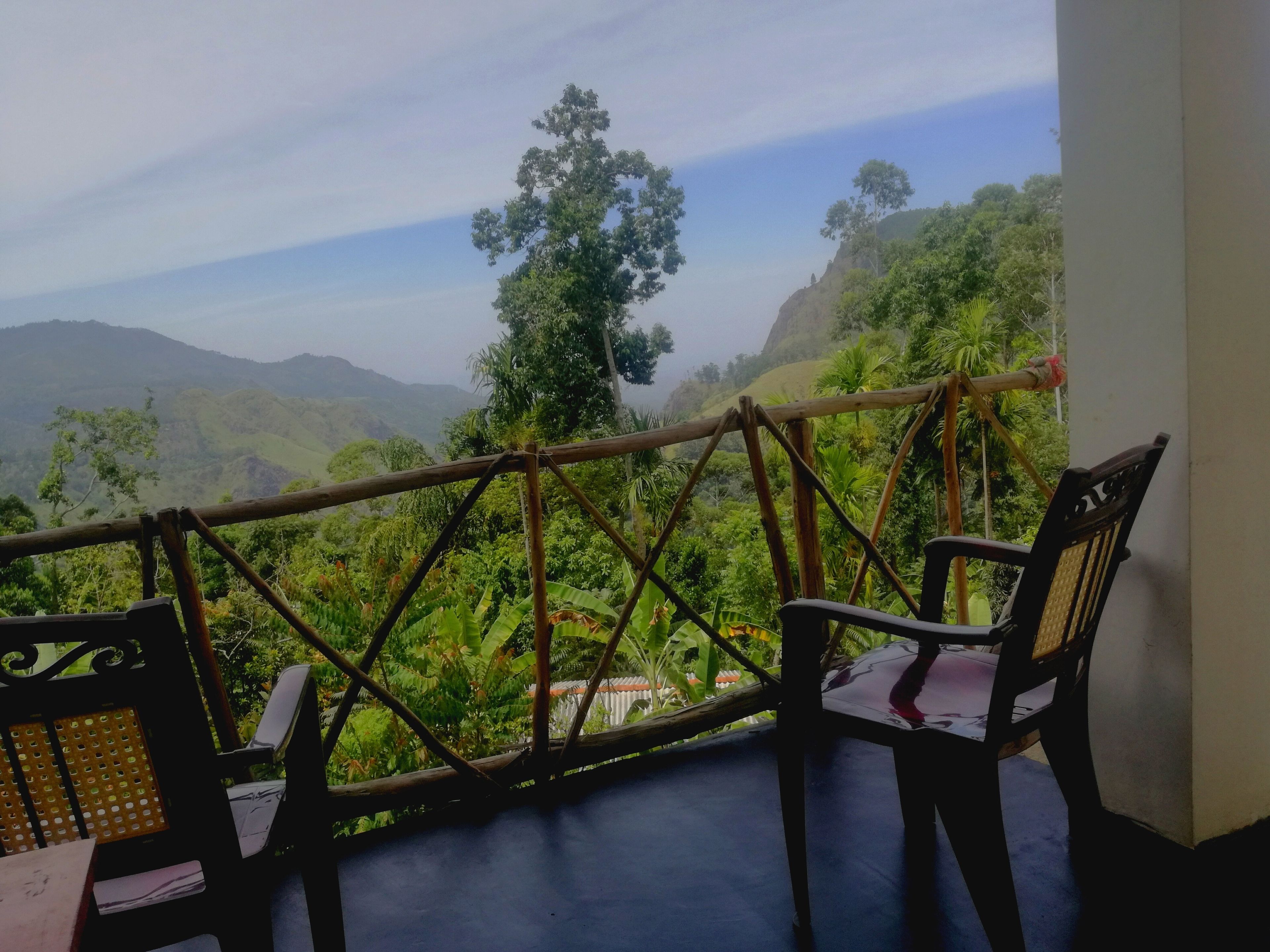 A balcony with a view of lush greenery and mountains. There are two chairs and a table on the balcony.