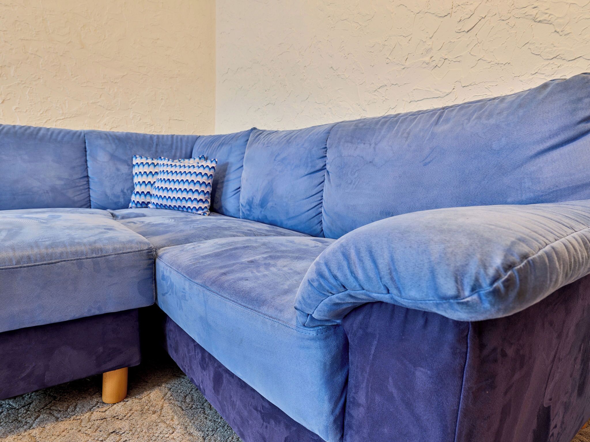 A blue sectional sofa with wooden legs, a patterned pillow, and a textured wall in the background.