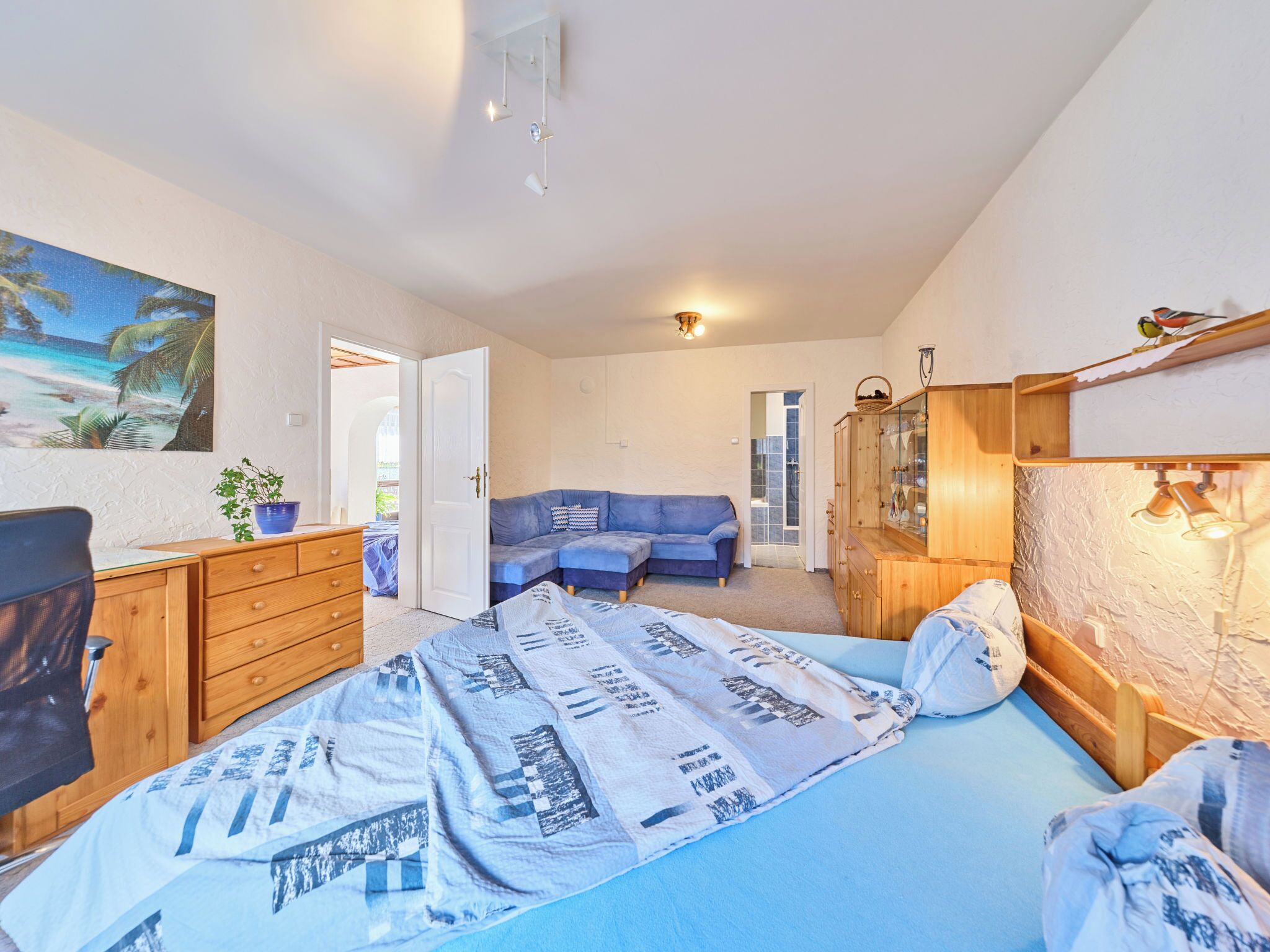 Un dormitorio con una cama, un sofá azul, un aparador de madera, un escritorio y un cuadro con una playa.