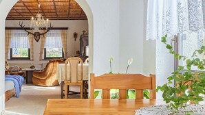 A cozy room with a wooden dining table, a comfortable sofa, and a view through an archway to another room with a chandelier and a deer head decoration.